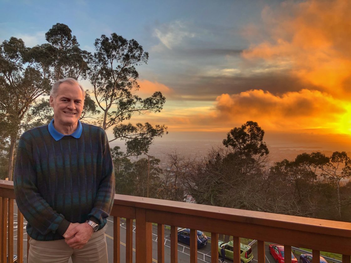 Steve Kevan wearing sweater, standing at a wood railing in front of a sunset