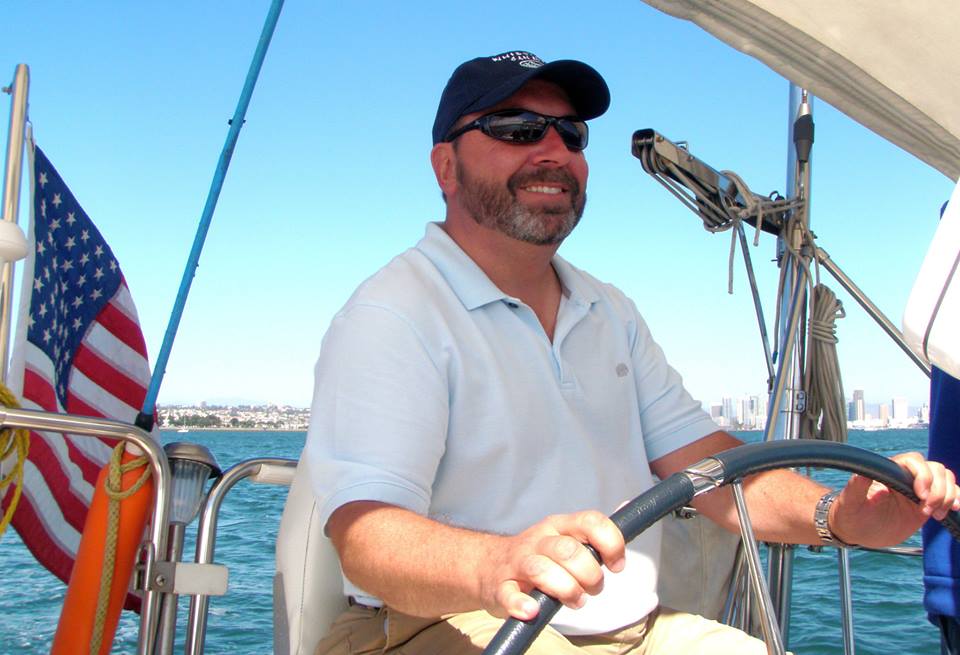 man wearing a baseball cap, sunglasses, and white polo shirt at the wheel of a boat on the water