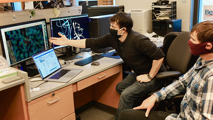 Photo of two masked individuals in an office setting looking at models on computer screens.