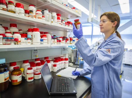 A woman in a lab coat holding a container of a chemical substance in front of a shelf of many containers.