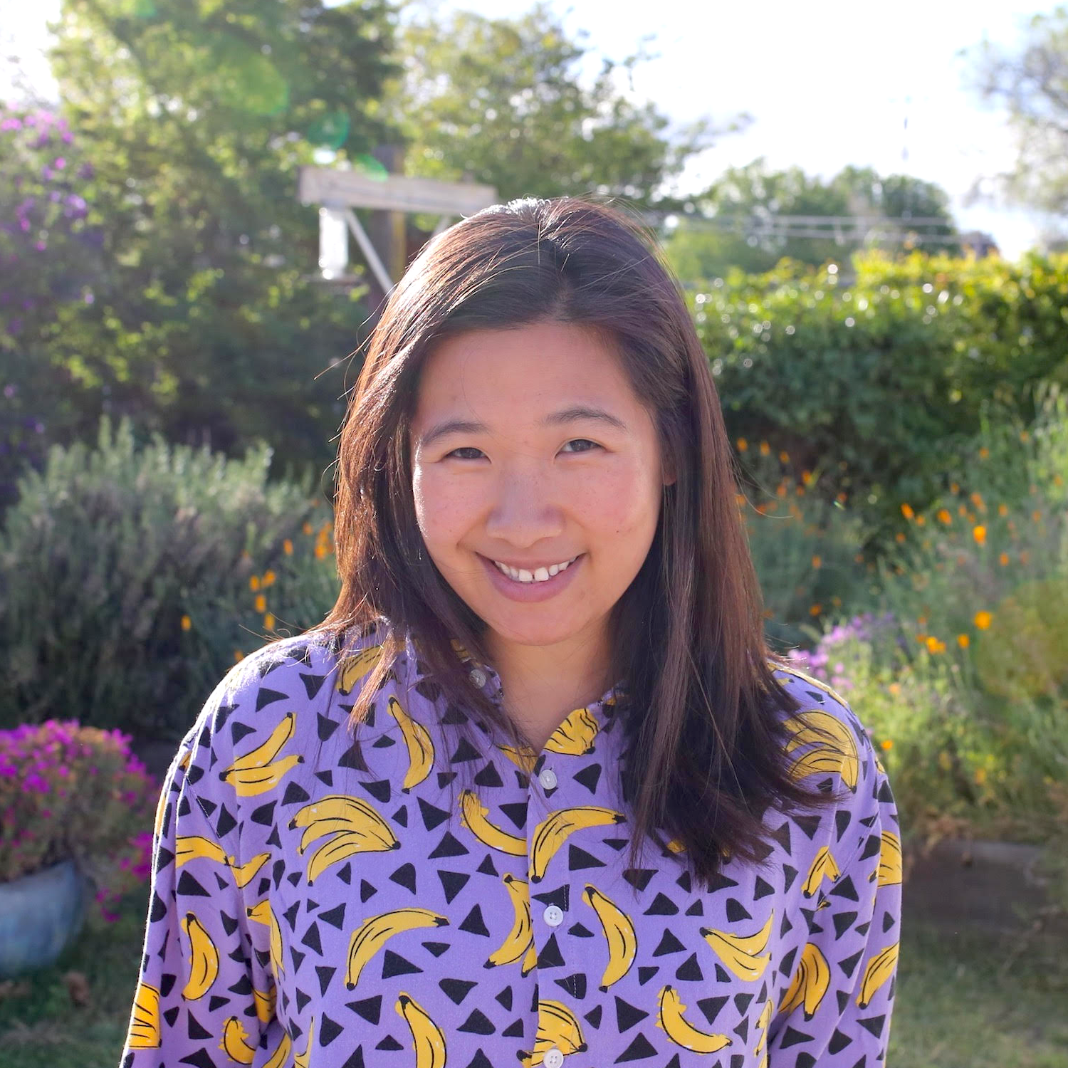 smiling woman wearing purple shirt with banana print in front of green plants