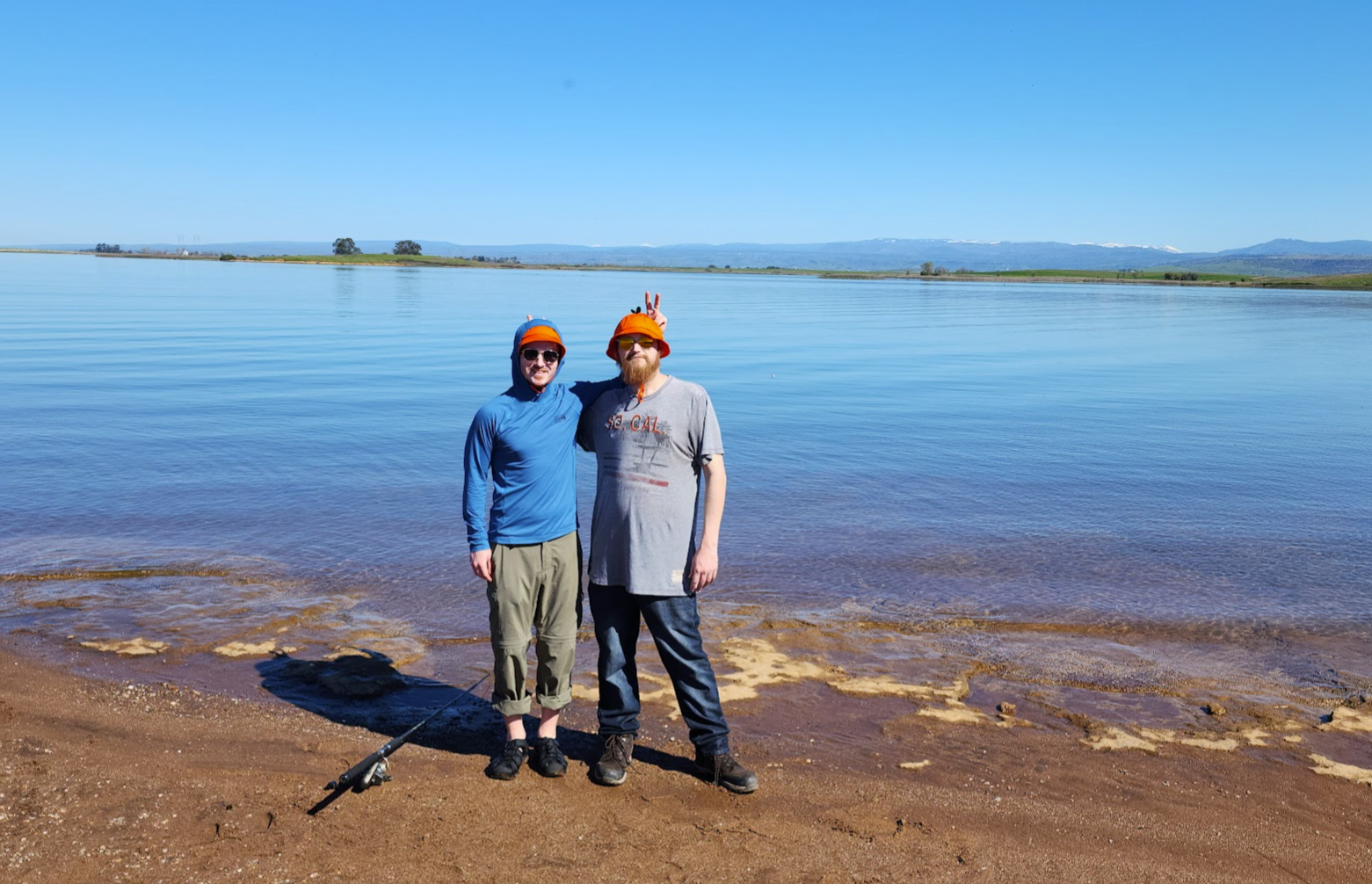 Two men standing in front of water with a fishing pole at their feet