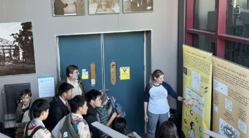 Female scientist giving a tour to middle school aged students and pointing at an illustration on a poster.