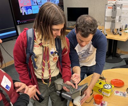 Teacher reaching over table and holding a scientific device and showing a student how to use the device.