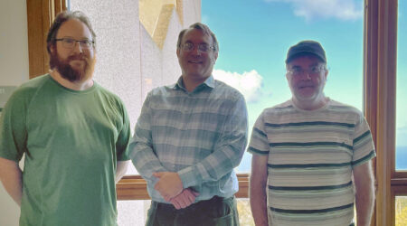 Snapshot of three men standing in front of a bright window with blue sky in the background.