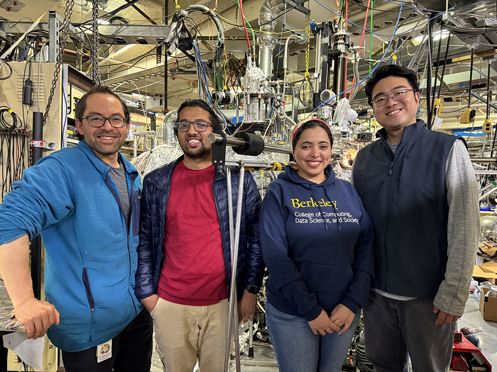 Photo of three men and one woman smiling and standing in front of complicated scientific equipment, covered in foil and strewn with chains and colored cables.