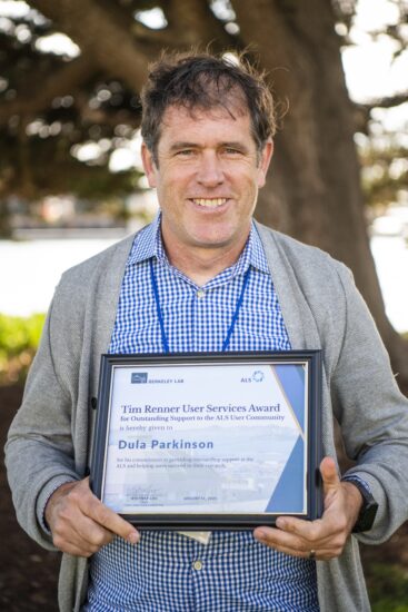 A man standing outdoors holding a framed awards certificate. He has a big smile on his face.