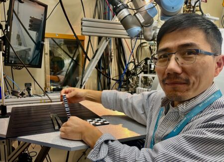 Person stands in front of a large instrument and holds up a metal bar against an elevated platform. A checkered square is visible in the corner of the platform.