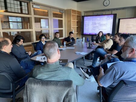 A group of people sitting around a table discussing with a big screen in the background.