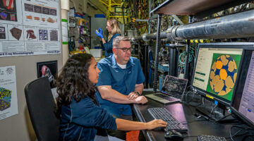 a man and woman sit at a computer screen, and a woman is standing in the background