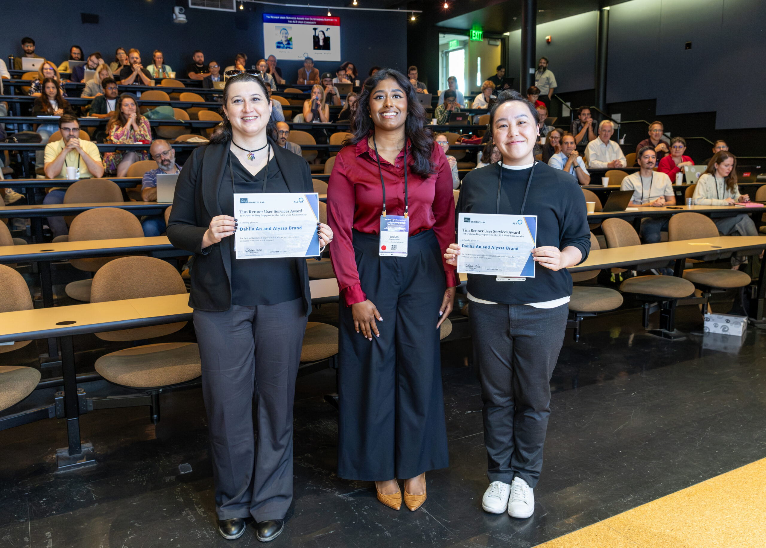 Three woman in an auditorium, two of them are holding certificates.