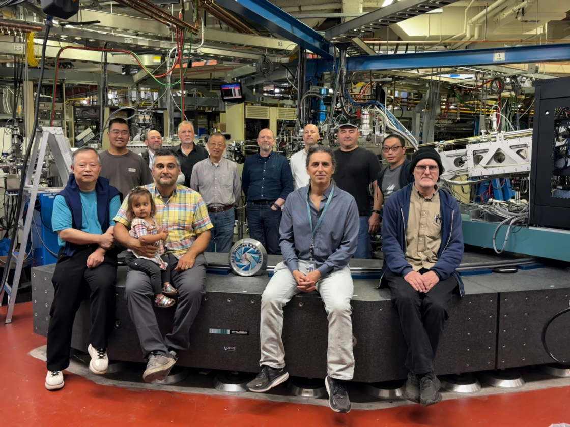 Group of eleven people posed on an instrument platform at a crowded beamline, with a round “ALS Beamline 6.0.2 QERLIN” plaque displayed in front.