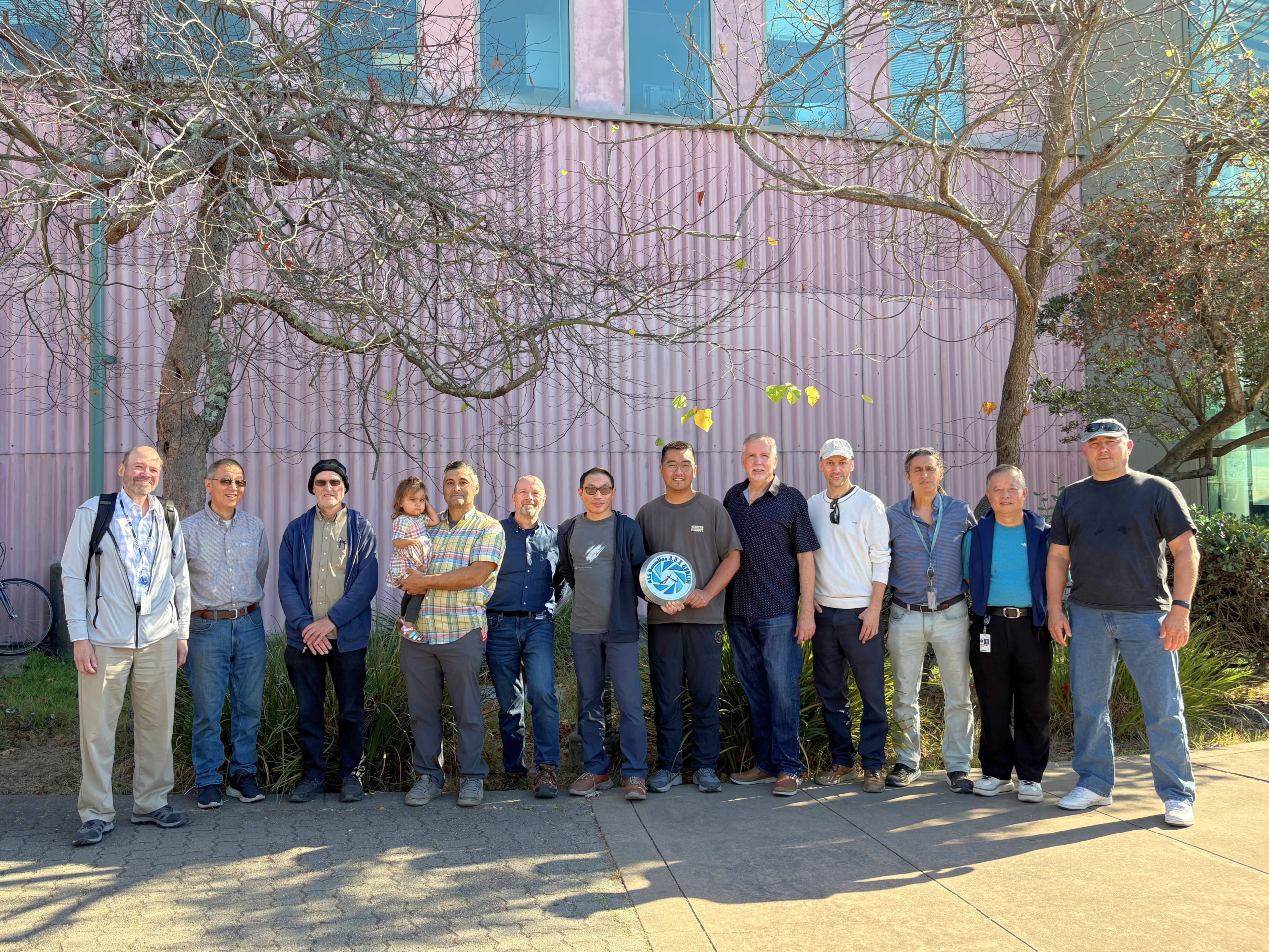 Group of eleven people stand in front of a pink building and two trees 