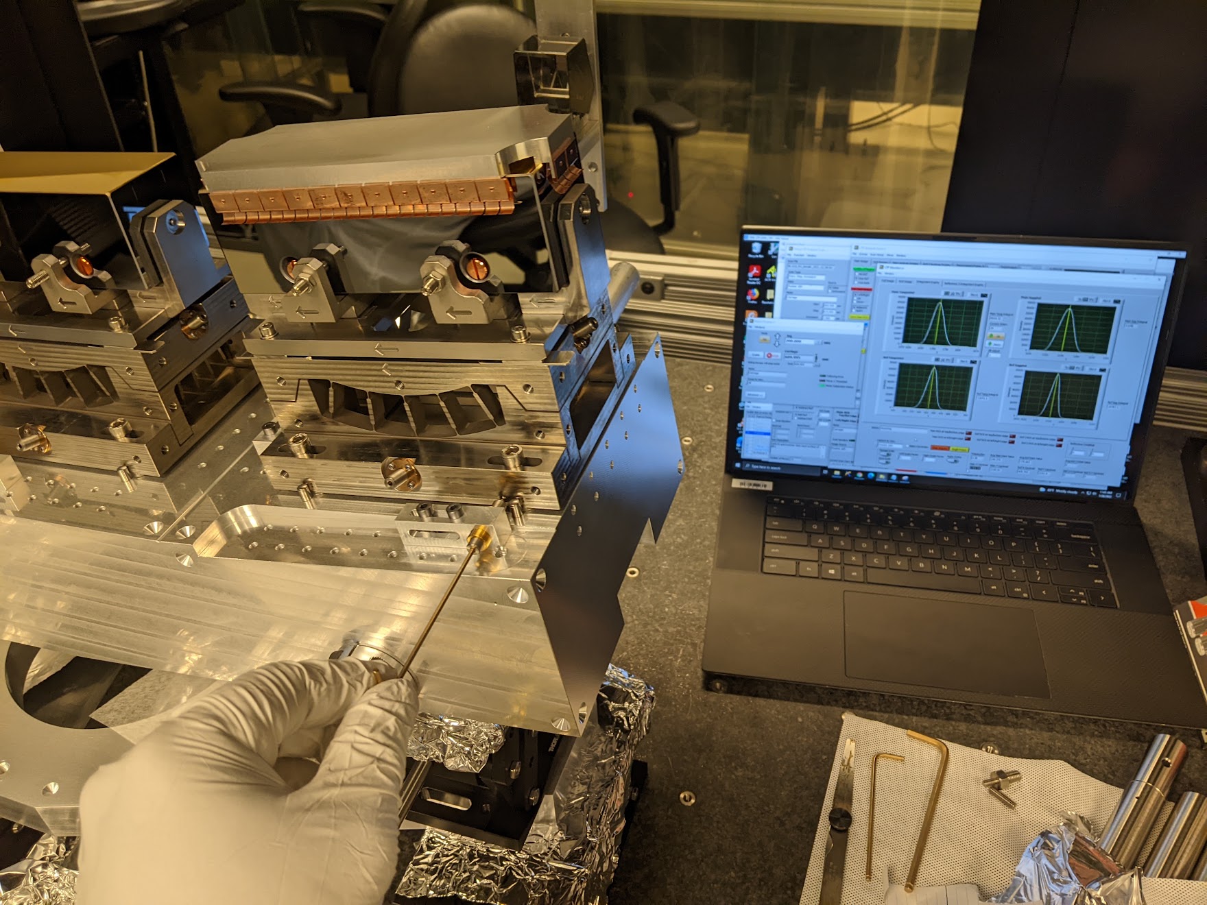 A gloved hand turns a dial on a scientific instrument with a feedback screen in the background
